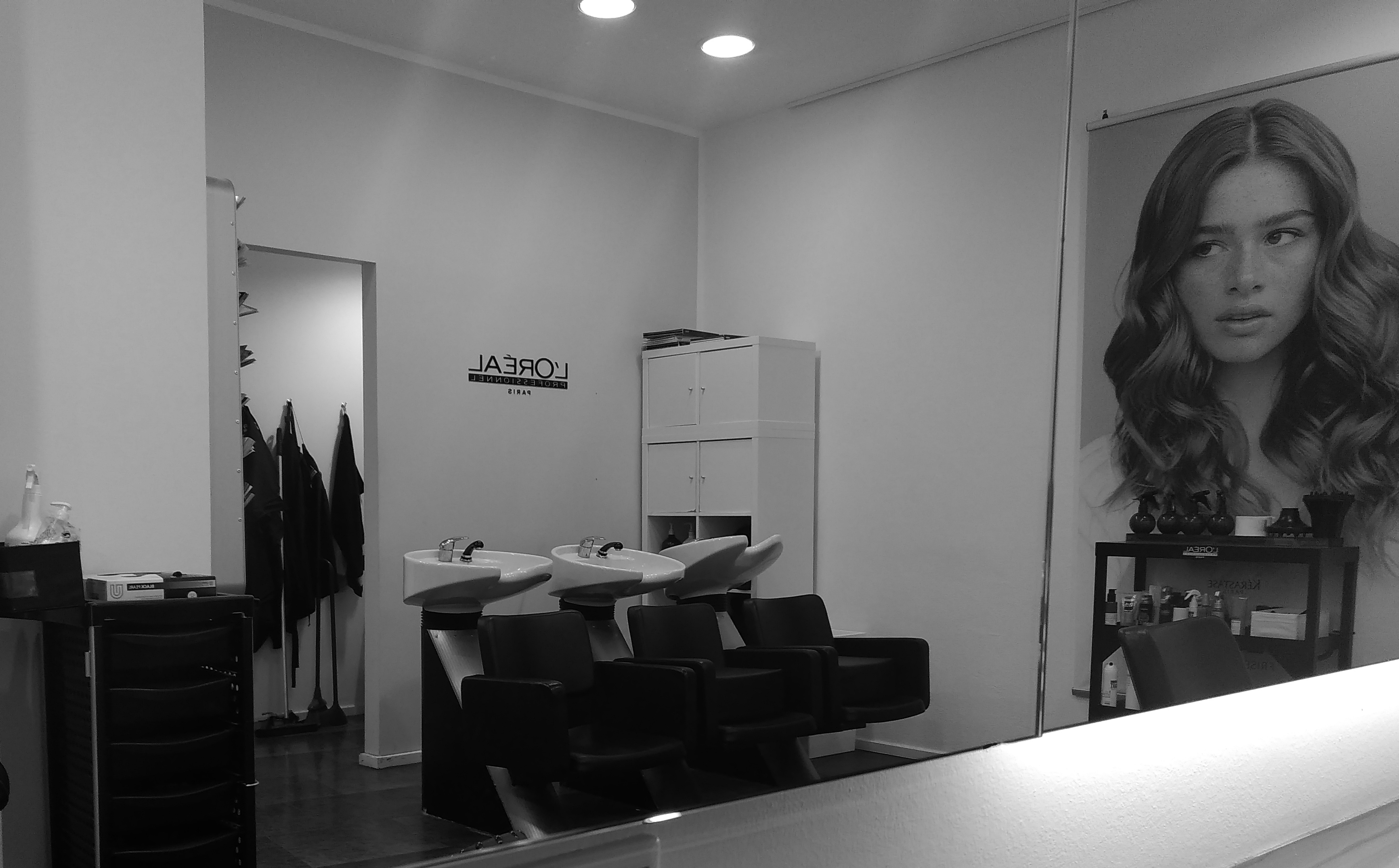 a black and white image of a hairdressing salon. There are two sinks in the back right hand corner and a poster on the wall which shows a headshot of a long haired woman. There is a doorway with some aprons hanging up.