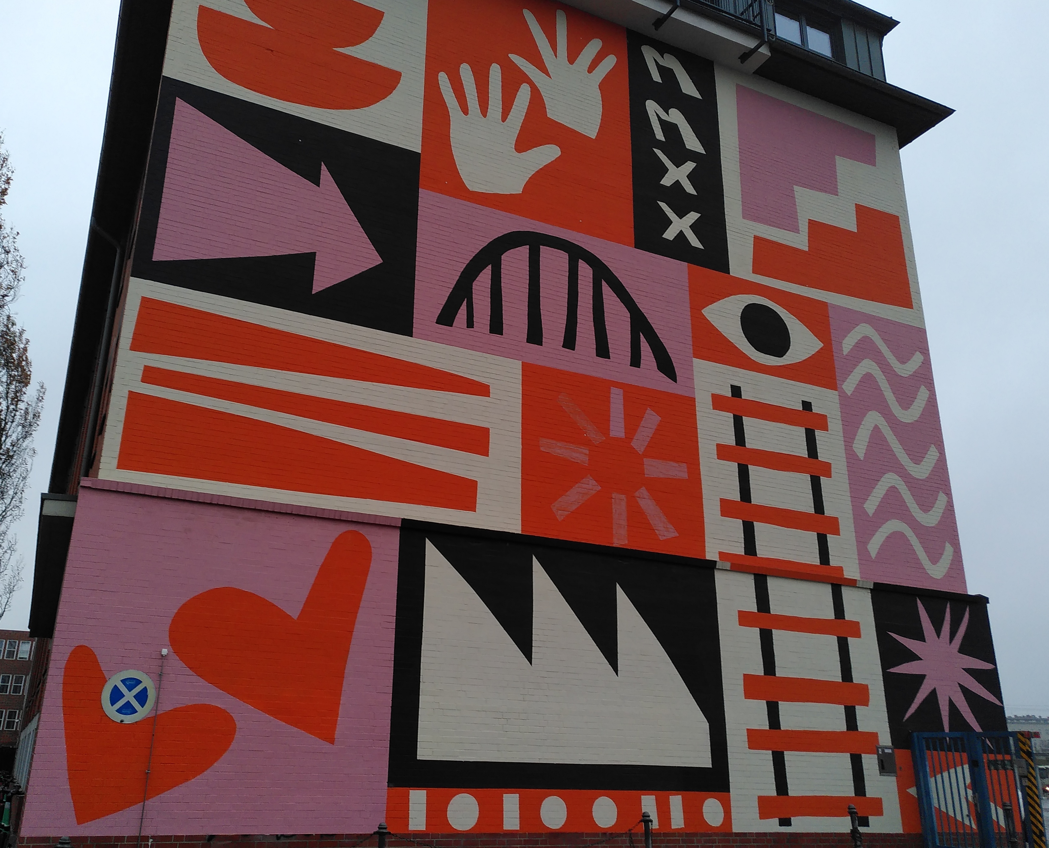 a painted mural on the side of a building. The images are in black, white, red and pink. from the top left: on a white background two half circles stacked, on a red background two white hands, on a blackbackground two letter m's and two letter x's, on a white background pink and orange stairs facing inwards towards each other, on a black background a pink arrow pointing right, on a pink background a black curved bridge, on a red background a white eye with black pupil, on a pink background white squiggly lines, on a white background thick red ramp lines, on a red background a outlines of sun rays in pink, on a white background black train tracks with red sleepers crossing them, on a black background a pink star, on a pink background two red love hearts, on a black background a white jagged outline like a mountain range, then a red backgroud with lines and dots.